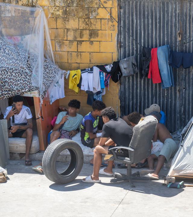 Jugendliche Migranten im spanischen Ceuta sitzen unter einer Wäscheleine auf Matratzen und in Zelten an einer Hauswand.