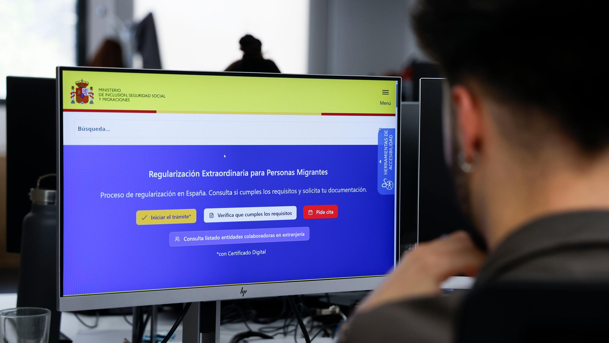 Ein junger Mann mit Ohrring schaut auf einen Computer. Darauf ist das gelb-blaue Online-Portal der spanischen Regierung zu sehen, über welches Migranten einen Aufenthaltstitel und eine Arbeitsgenemigung beantragen können.