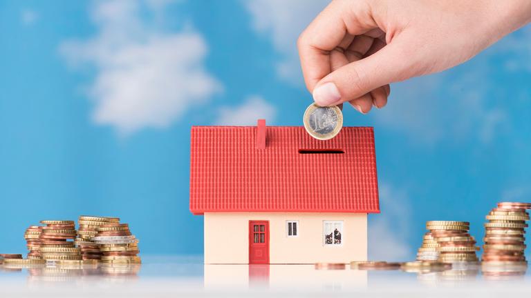 Eine Hand steckt Geld in eine Spardose in Form eines Hauses.