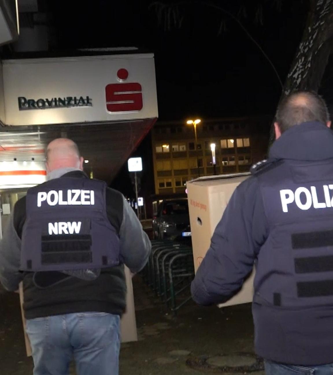 Einsatzkräfte der Polizei verlassen nach der Durchsuchung die Sparkassenfiliale mit Beweismaterial. 