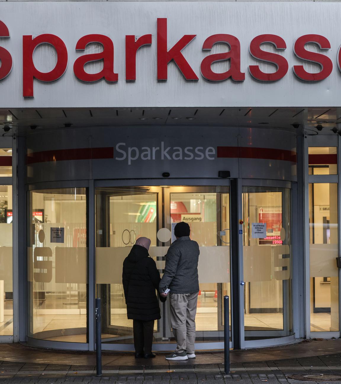 Zwei Passanten, die von hinten zu sehen sind, stehen vor einer geschlossenen Sparkasse.