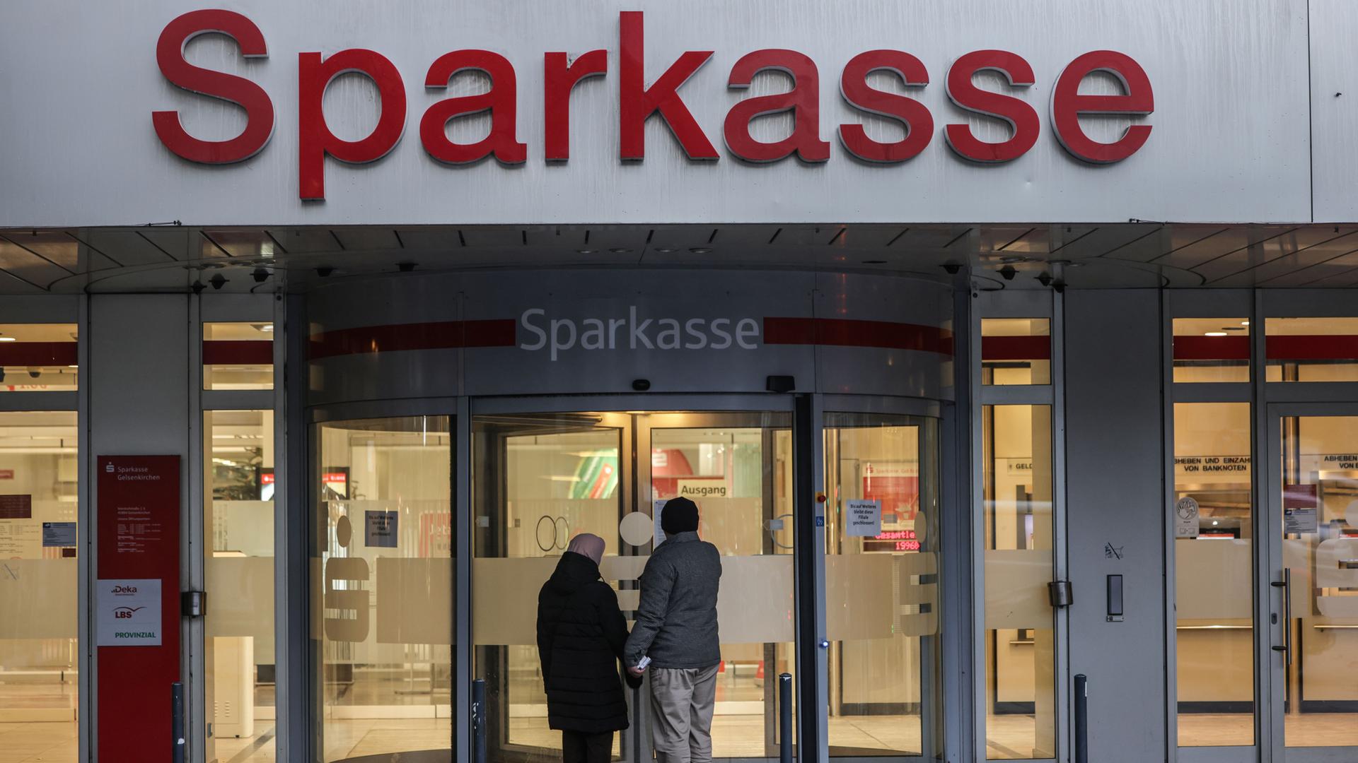 Zwei Passanten, die von hinten zu sehen sind, stehen vor einer geschlossenen Sparkasse.