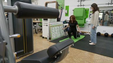 In einem Fitnessstudio geht eine Frau mit Multipler Sklerose auf ein Knie herunter, während eine andere Frau neben ihr steht.