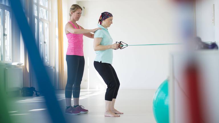 Eine Sporttherapeutin steht hinter einer Krebspatientin, die eine Übung mit Bändern ausführt.