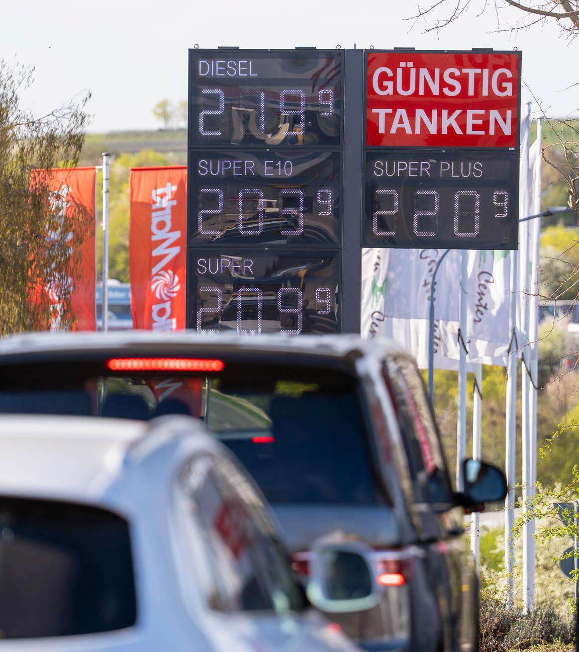 "Günstig Tanken" steht auf einer Anzeigetafel mit den Spritpreisen an einer Tankstelle im Landkreis Alzey-Worms, davor reihen sich Autos aneinander.