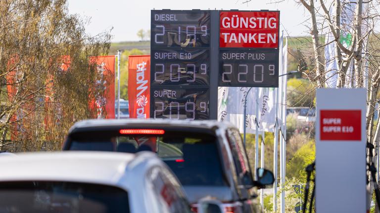 "Günstig Tanken" steht auf einer Anzeigetafel mit den Spritpreisen an einer Tankstelle im Landkreis Alzey-Worms, davor reihen sich Autos aneinander.