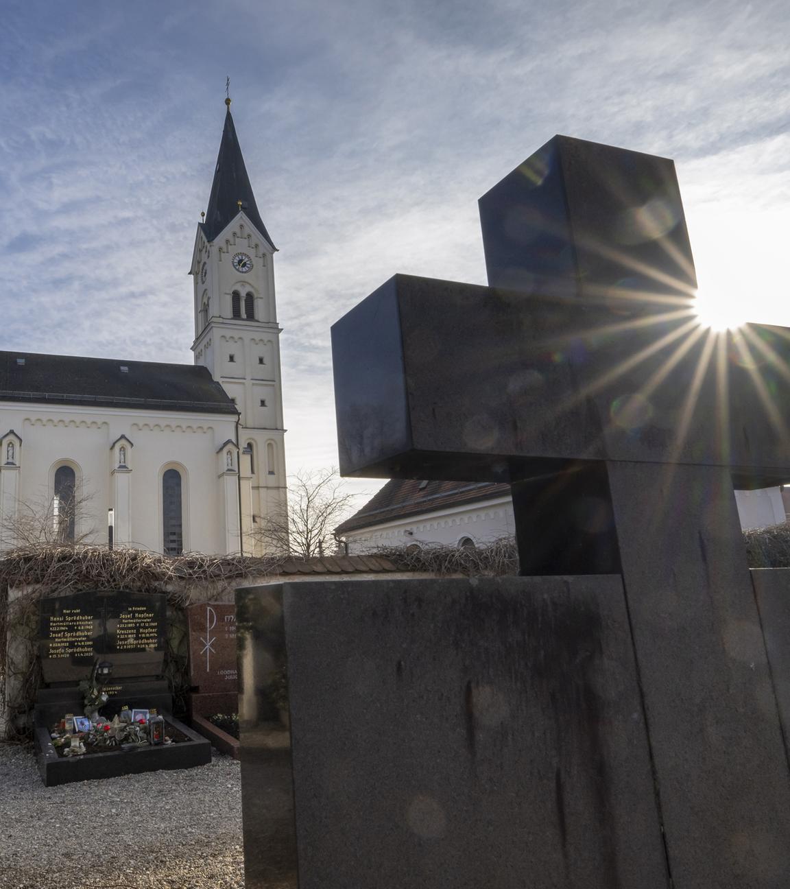 Das Kreuz eines Grabsteins ragt vor der katholische Kirche St. Nikolaus in Garching An Der Alz in den Himmel.