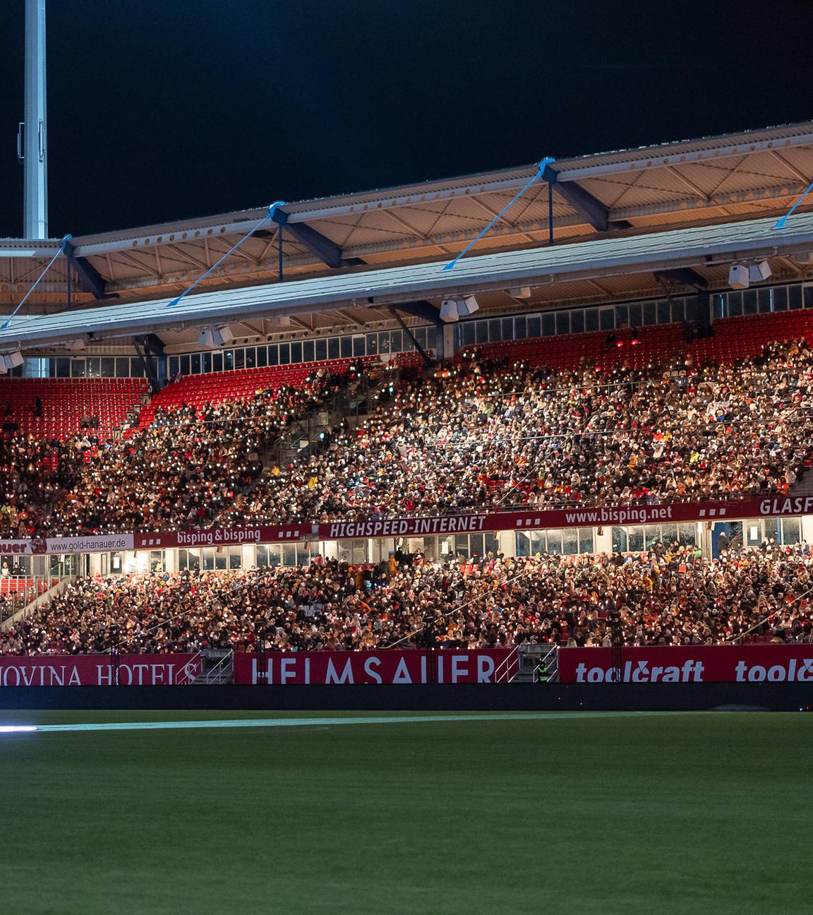  Die Tribünen des Max-Morlock-Stadions sind bis auf den letzten Platz gefüllt. Tausende Besucher:innen sitzen mit Kerzen in der Hand und schaffen gemeinsam ein beeindruckendes Lichtermeer beim Großen Nürnberger Rote Engel Adventssingen. 