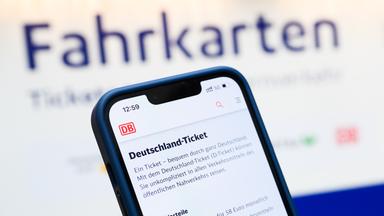 Eine Person hält am Hauptbahnhof ein Smartphone in der Hand, auf dessen Display eine Information zum Kauf des Deutschlandtickets zu sehen ist, während sie vor einem Fahrkartenautomaten steht. 