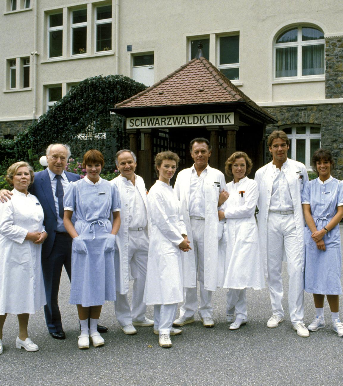 "Die Schwarzwaldklinik - Die Heimkehr (1)": Oberschwester Hildegard (Eva Maria Bauer), Verwaltungsdirektor Alfred Mühlmann (Alf Marholm), Schwester Ina (Gaby Fischer), Dr. Gerd Wolter (Franz Rudnick), Dr. Katarina Gessner (Ilona Grübel), Prof. Klaus Brinkmann (Klausjürgen Wussow), Schwester Christa (Gaby Dohm), Dr. Udo Brinkmann (Sascha Hehn) und Schwester Elke (Barbara Wussow) stehen nebeneinander vor der Schwarzwaldklinik und lächeln Richtung Kamera.
