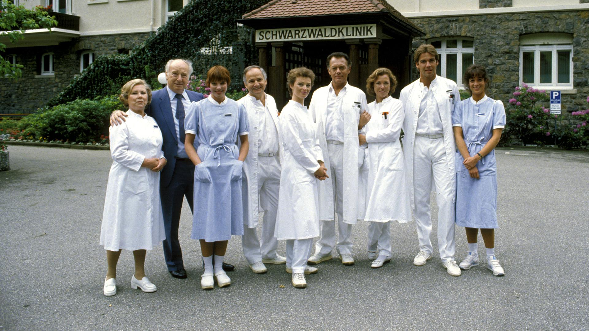 "Die Schwarzwaldklinik - Die Heimkehr (1)": Oberschwester Hildegard (Eva Maria Bauer), Verwaltungsdirektor Alfred Mühlmann (Alf Marholm), Schwester Ina (Gaby Fischer), Dr. Gerd Wolter (Franz Rudnick), Dr. Katarina Gessner (Ilona Grübel), Prof. Klaus Brinkmann (Klausjürgen Wussow), Schwester Christa (Gaby Dohm), Dr. Udo Brinkmann (Sascha Hehn) und Schwester Elke (Barbara Wussow) stehen nebeneinander vor der Schwarzwaldklinik und lächeln Richtung Kamera.