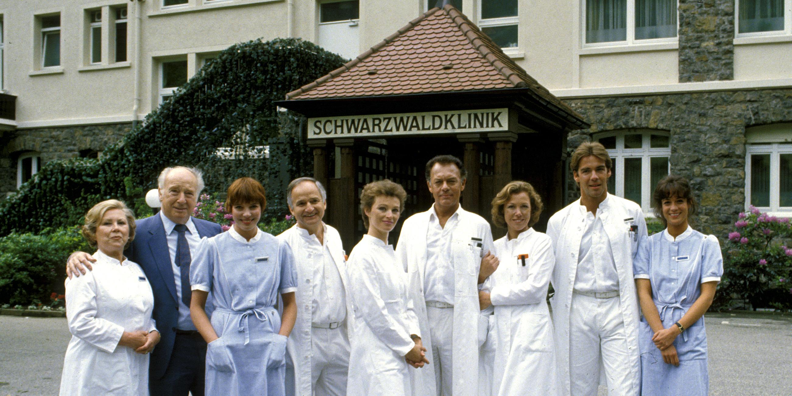 "Die Schwarzwaldklinik - Die Heimkehr (1)": Oberschwester Hildegard (Eva Maria Bauer), Verwaltungsdirektor Alfred Mühlmann (Alf Marholm), Schwester Ina (Gaby Fischer), Dr. Gerd Wolter (Franz Rudnick), Dr. Katarina Gessner (Ilona Grübel), Prof. Klaus Brinkmann (Klausjürgen Wussow), Schwester Christa (Gaby Dohm), Dr. Udo Brinkmann (Sascha Hehn) und Schwester Elke (Barbara Wussow) stehen nebeneinander vor der Schwarzwaldklinik und lächeln Richtung Kamera.