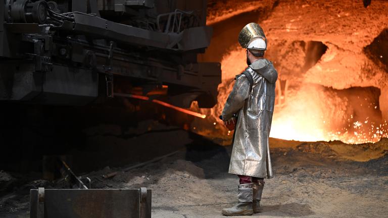 Stahlarbeiter steht an einem Hochofen, im Hintergrund leuchtet es rot durch die Glut/Hitze