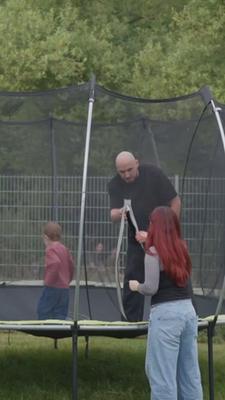 Ein Mann mit einem kleinen Kind auf einem Trampolin. Eine Frau steht vor dem Trampolin