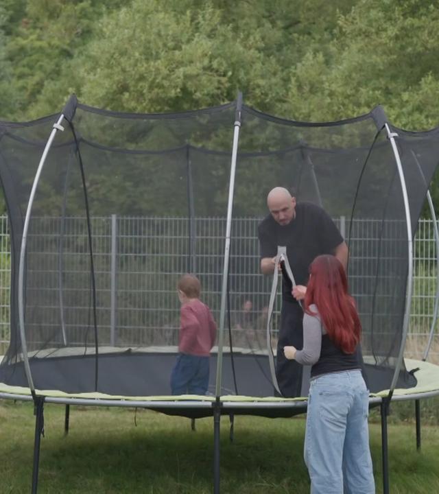 Ein Mann mit einem kleinen Kind auf einem Trampolin. Eine Frau steht vor dem Trampolin