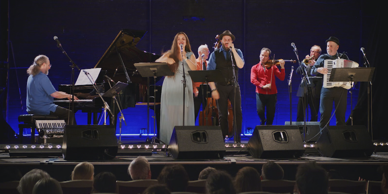 Standbild aus dem Dokumentarfilm "I dance but my heart is crying": Mehrere Menschen stehen vor Publikum auf einer Bühne und machen Musik