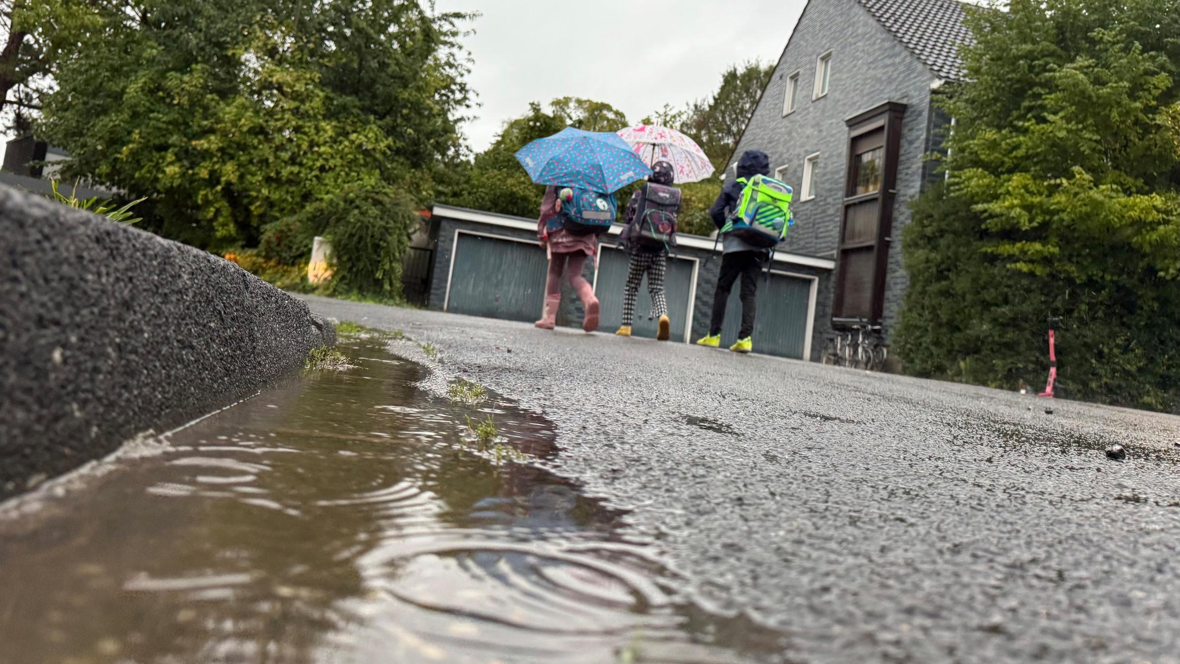 Drei Schulkinder gehen bei Regenwetter durch ein Wohngebiet zur Schule