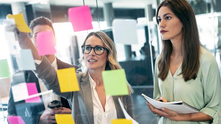 Geschäftsfrau diskutiert über Klebezettel an der Glaswand, während sie mit einer Kollegin im Büro steht