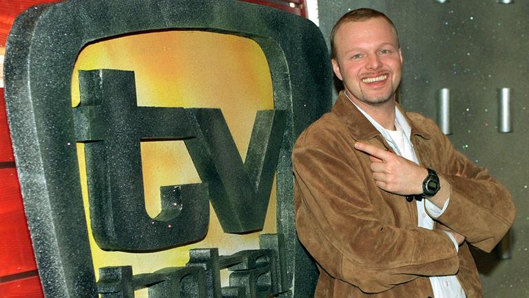 Stefan Raab steht vor einem gropen "TV total"-Schild und lächelt in die Kamera