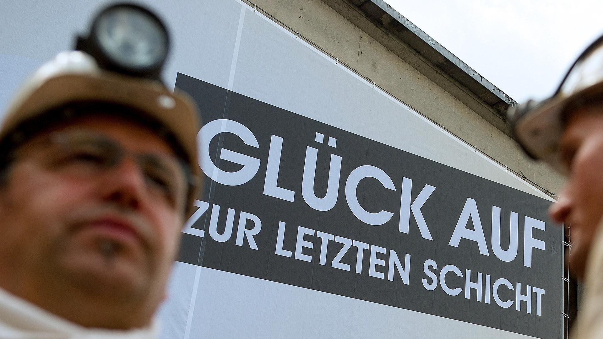 Archiv: Zwei Bergmänner vor einem Plakat mit der Aufschrift "Glück auf zur letzten Schicht"