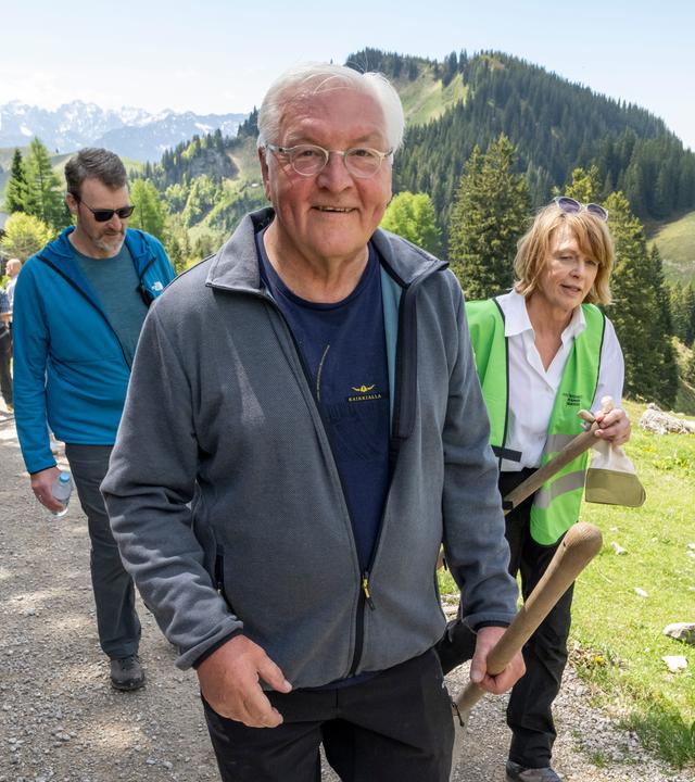Bundespräsident Frank-Walter Steinmeier und seine Frau Elke Büdenbender mit Schaufeln in der Hand vor Bergkulisse.