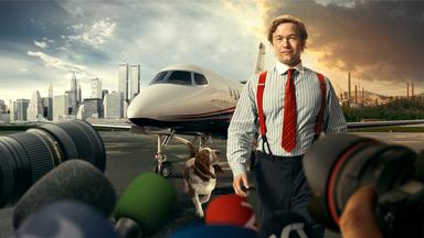 Stenbeck steht, in einem gestreiften Hemd mit roter Krawatte und roten Hosenträgern, auf einem Flugfeld. Sein Hund, ein braun-weißer Basset, schaut von links unten hoch zu ihm. Direkt hinter ihm steht ein moderner Privat-Jet. Im Hintergrund sieht man eine geteilte Landschaft, links die New Yorker Skyline, rechts eine ländliche Landschaft mit einer rauchenden Fabrik darauf. Der Himmel über New York ist weitgehend klar und blau, über der Fabrik ziehen dunkle, rötlich gefärbte Wolken. Im Vordergrund sind Kameras und Mikrofone aufgebaut.