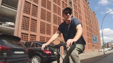 Stephan Kampwirth fährt auf einem Fahrrad eine Straße entlang. Er trägt eine dunkle Kappe, eine Sonnenbrille, ein dunkles T-Shirt und eine helle Hose. Beide Hände liegen am Lenker. 