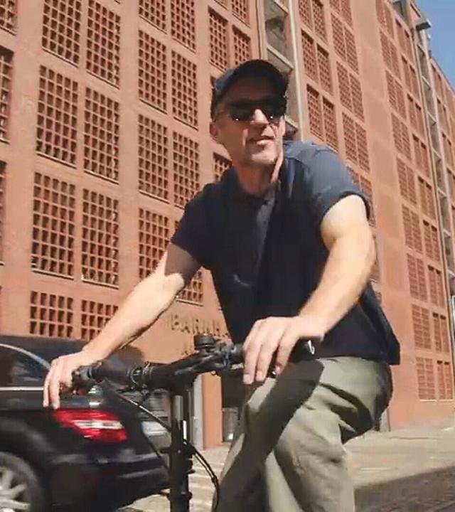 Stephan Kampwirth fährt auf einem Fahrrad eine Straße entlang. Er trägt eine dunkle Kappe, eine Sonnenbrille, ein dunkles T-Shirt und eine helle Hose. Beide Hände liegen am Lenker. 