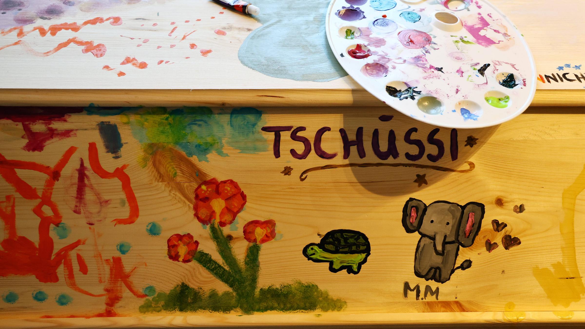 Ein schlichter Holzsarg wurde mit bunten Bildern bemalt. Eine Farbpalette liegt auf dem Deckel.