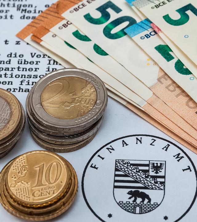 Zu sehen ist ein Bescheid, der vom Finanzamt abgestempelt ist, auf dem Geldmünzen und Geldscheine liegen.