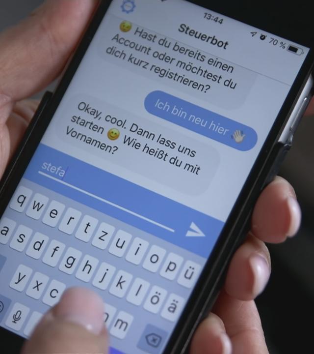 Ein Handy in einer Hand. Auf dem Handy ist ein Chat mit dem "Steuerbot" offen.