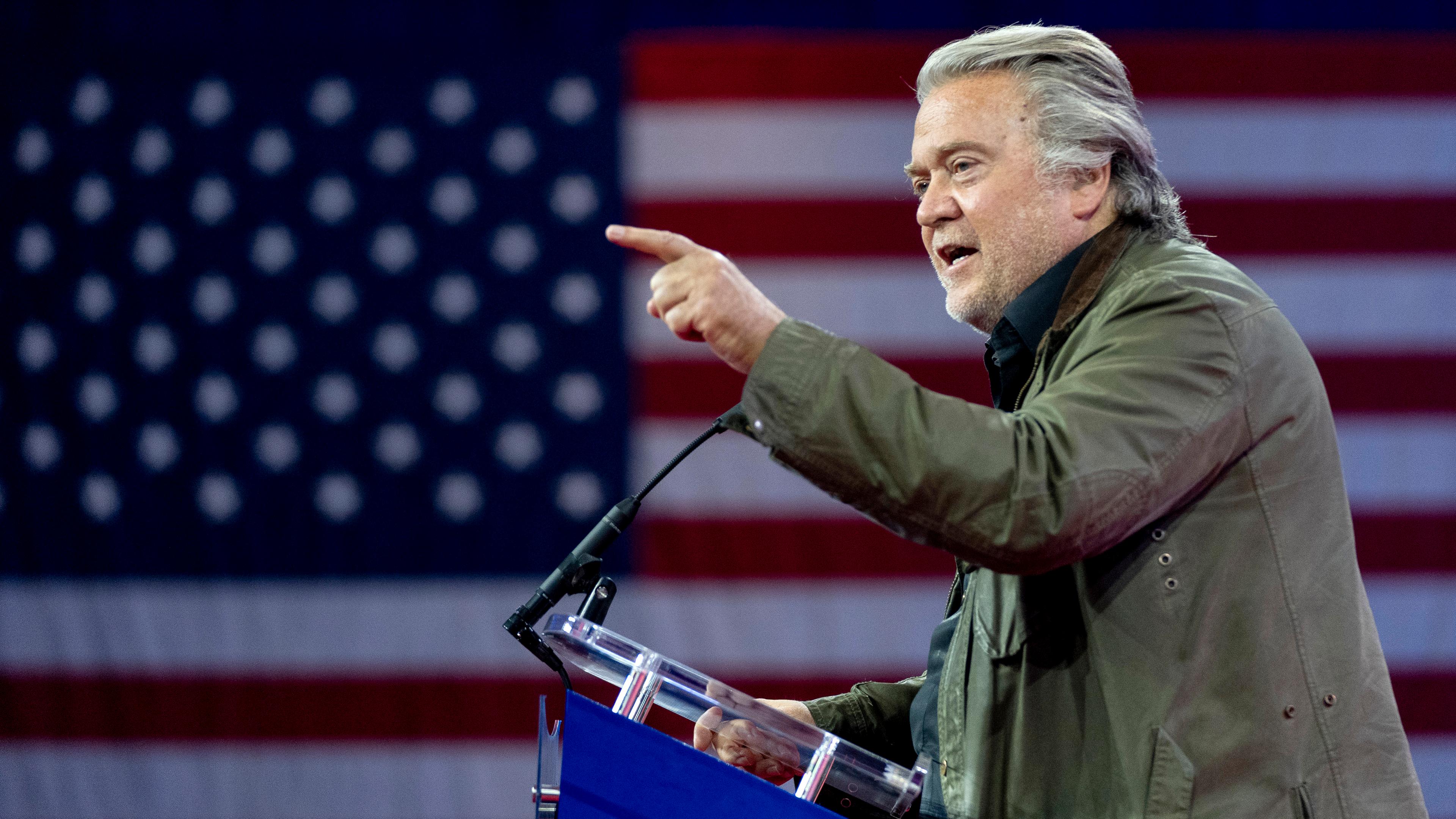 Steve Bannon steht auf einer Bühne am Punkt und spricht mit erhobenem Zeigefinger - im Hintergrund ist eine große US-Flagge zu sehen.