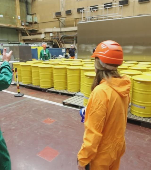 Reporterin steht in Schutzkleidung vor gelben Fässern