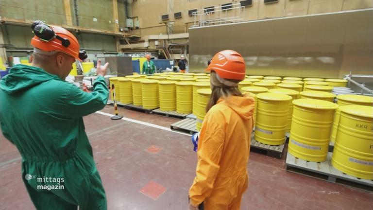 Reporterin steht in Schutzkleidung vor gelben Fässern