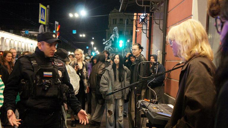 Eine junge blonde Frau bei einem Straßenkonzert, ein russischer Polizist steht ihr gegenüber