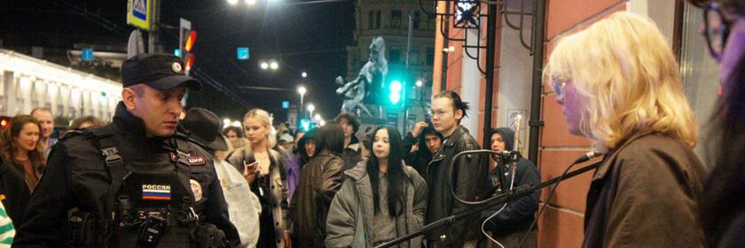 Eine junge blonde Frau bei einem Straßenkonzert, ein russischer Polizist steht ihr gegenüber