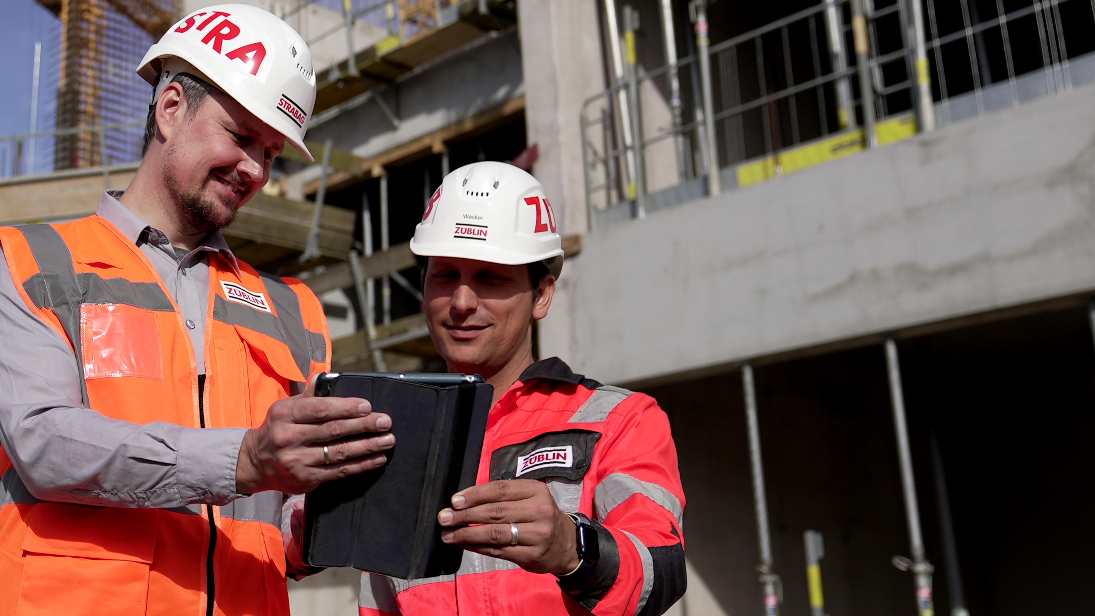Zwei Personen mit weißem Helm und orangener Weste stehen auf einer Baustelle und betrachten ein Tablet.