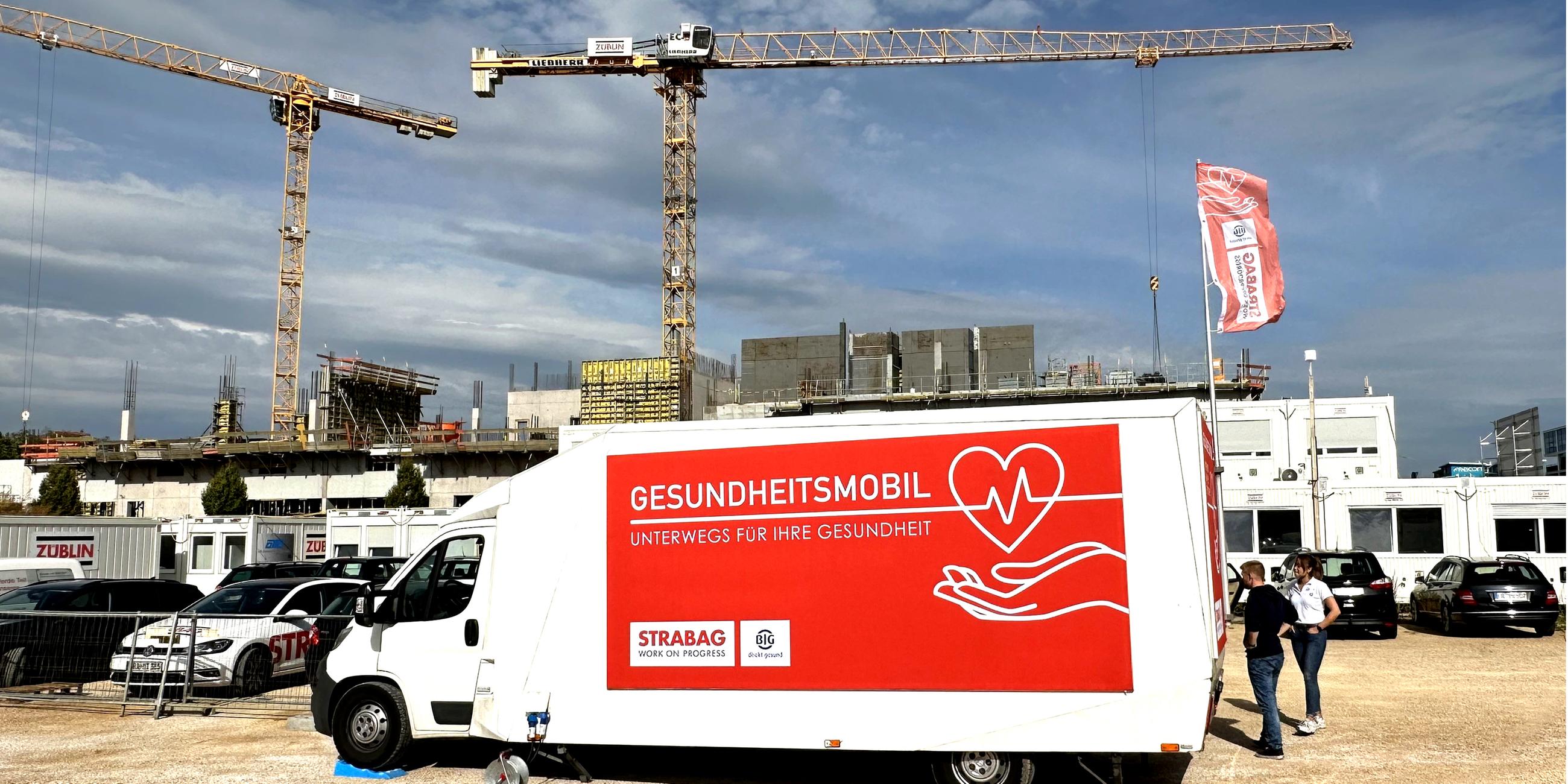 Ein kleiner Transportwagen steht auf einer Baustelle, daneben zwei Personen. Auf dem Wagen steht "Gesundheitsmobil - Unterwegs für Ihre Gesundheit".
