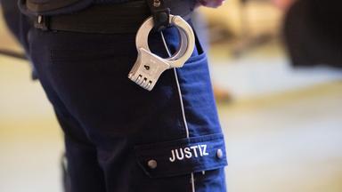 Symbolbild: Ein Justizbeamter mit Handschellen am Gürtel steht im Verhandlungssaal eines Verwaltungsgerichts in Nordrhein-Westfalen
