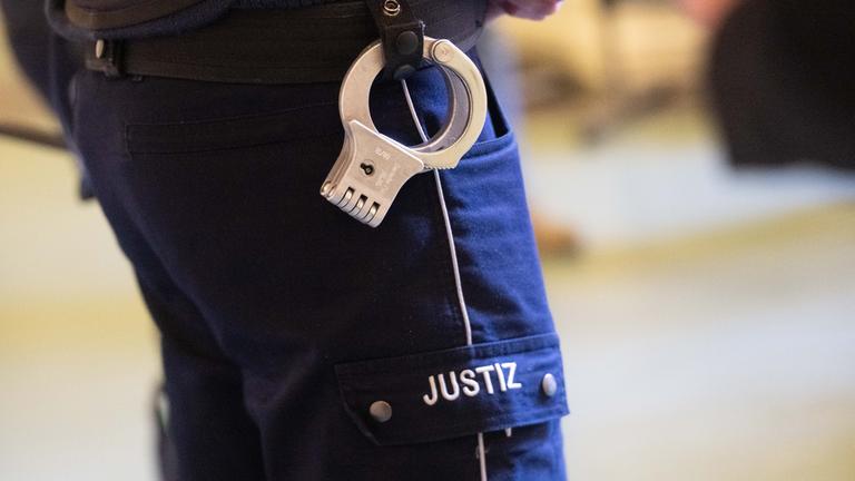 Symbolbild: Ein Justizbeamter mit Handschellen am Gürtel steht im Verhandlungssaal eines Verwaltungsgerichts in Nordrhein-Westfalen