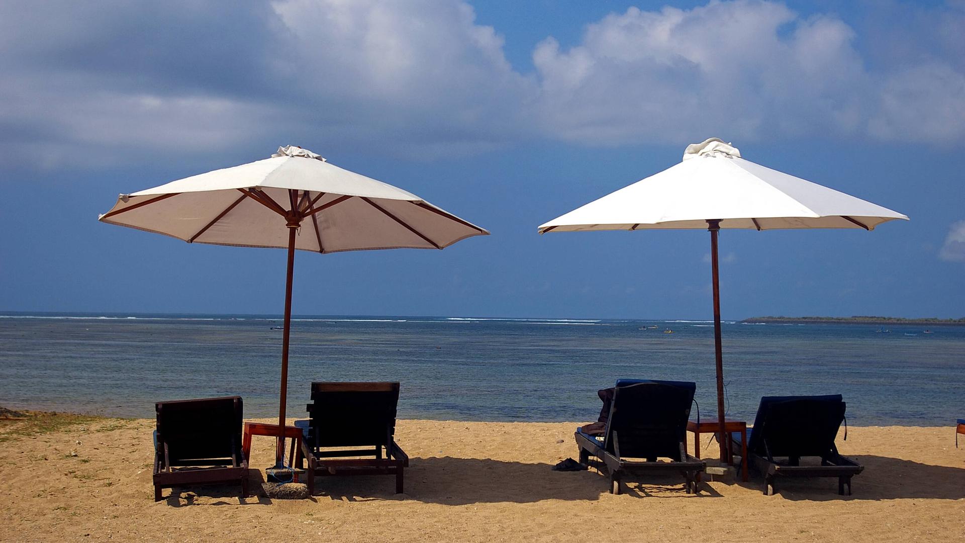 Zwei leere Strandliegen mit Sonnenschirmen stehen an einem Strand mit Blick auf das Meer.