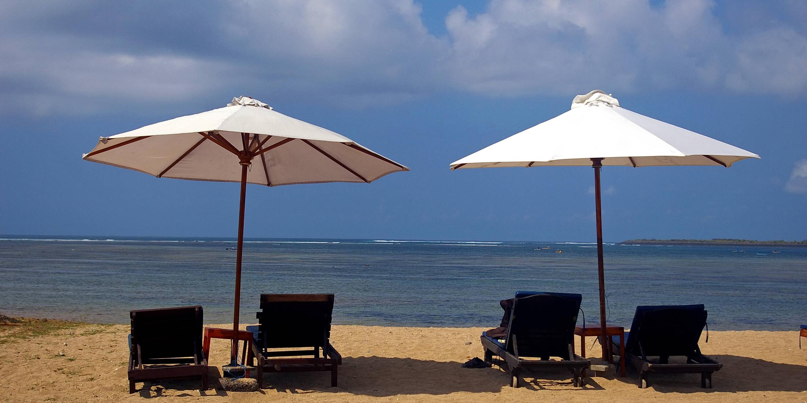 Zwei leere Strandliegen mit Sonnenschirmen stehen an einem Strand mit Blick auf das Meer.