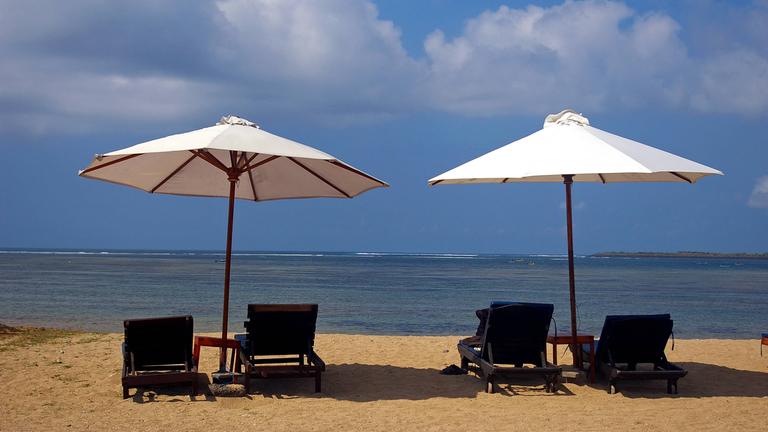 Zwei leere Strandliegen mit Sonnenschirmen stehen an einem Strand mit Blick auf das Meer.