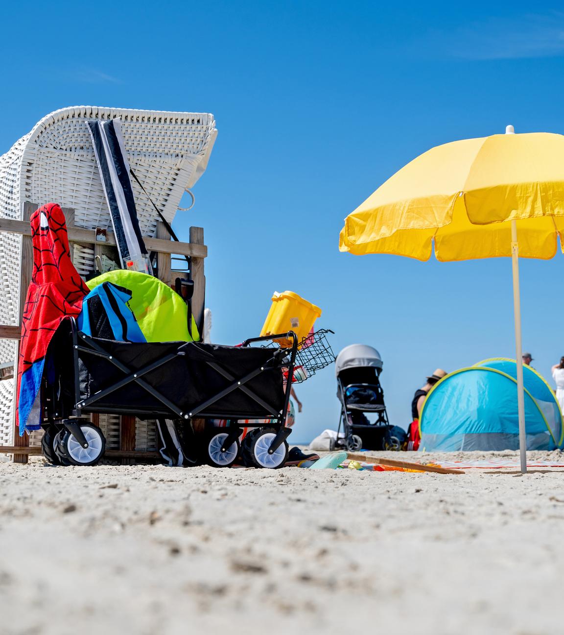 EIn Strandkorb, ein Sonnenschirm und ein Bollerwagen, im Hintergrund eine Strandmuschel und ein Kinderwagen, an einem Strand