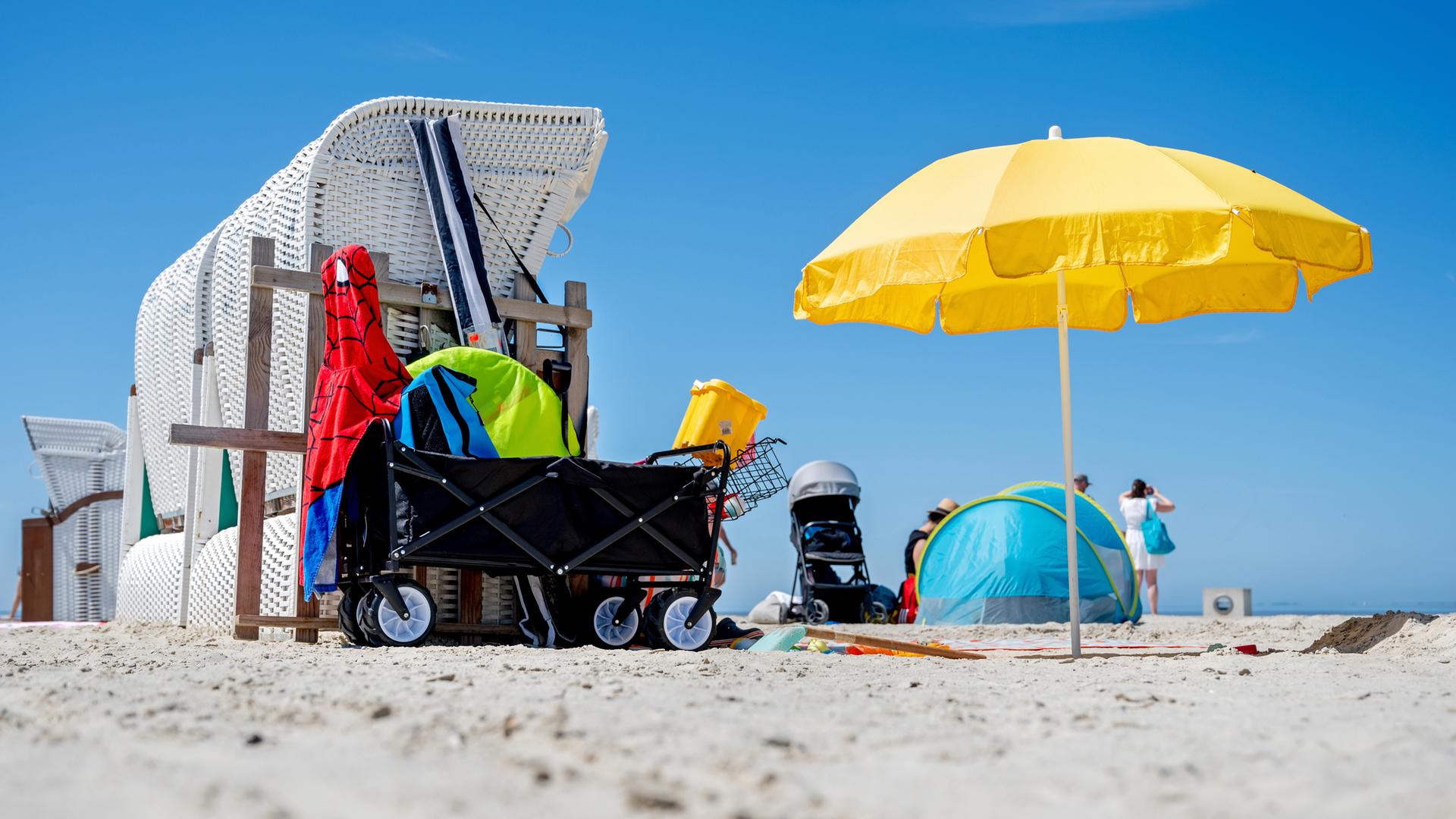 EIn Strandkorb, ein Sonnenschirm und ein Bollerwagen, im Hintergrund eine Strandmuschel und ein Kinderwagen, an einem Strand