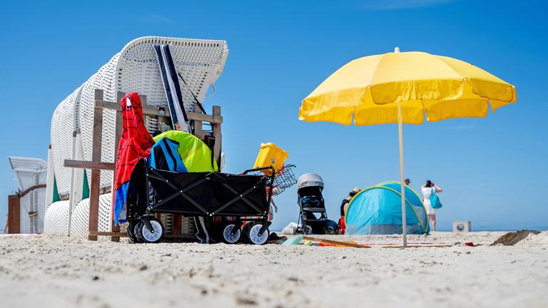 EIn Strandkorb, ein Sonnenschirm und ein Bollerwagen, im Hintergrund eine Strandmuschel und ein Kinderwagen, an einem Strand