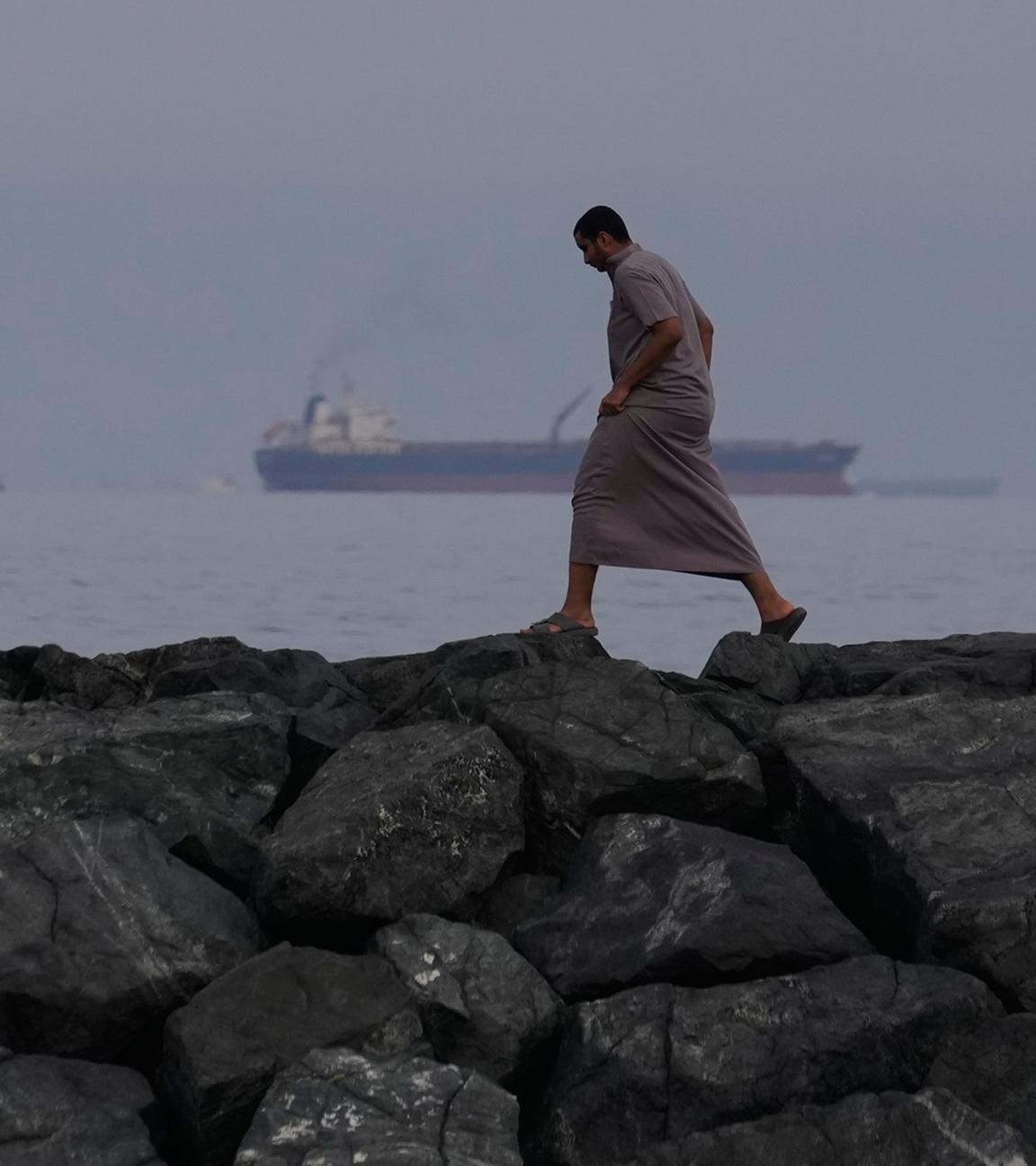 Ein Mann geht auf den Felsen am Ufer spazieren, während Öltanker und Frachtschiffe in der Straße von Hormus auf durchfahrt warten.