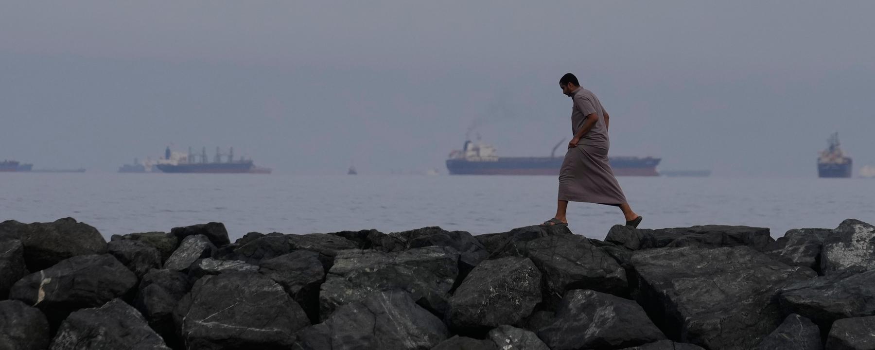 Ein Mann geht auf den Felsen am Ufer spazieren, während Öltanker und Frachtschiffe in der Straße von Hormus auf durchfahrt warten. (Archiv)