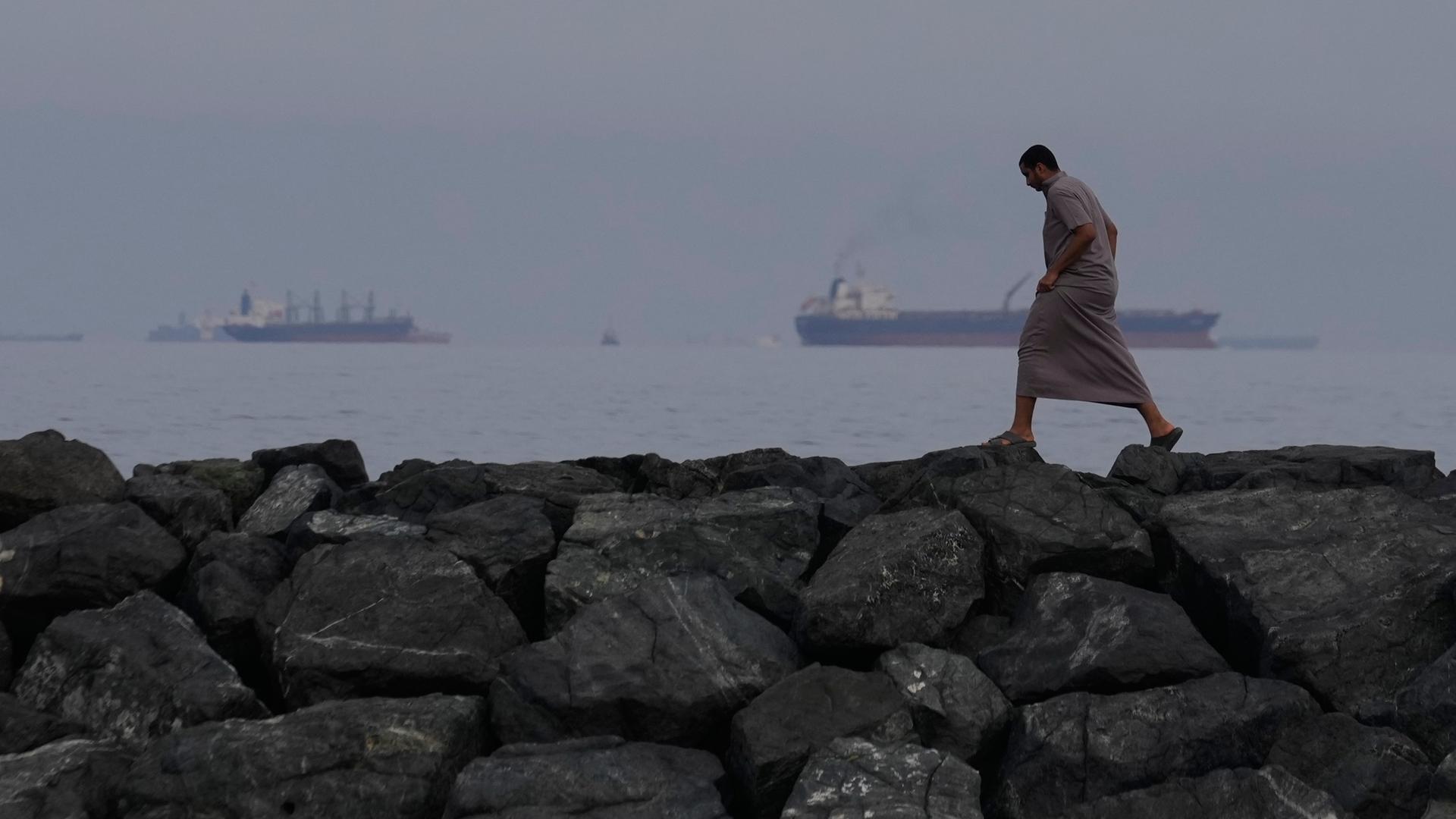 Ein Mann geht auf den Felsen am Ufer spazieren, während Öltanker und Frachtschiffe in der Straße von Hormus auf durchfahrt warten. (Archiv)