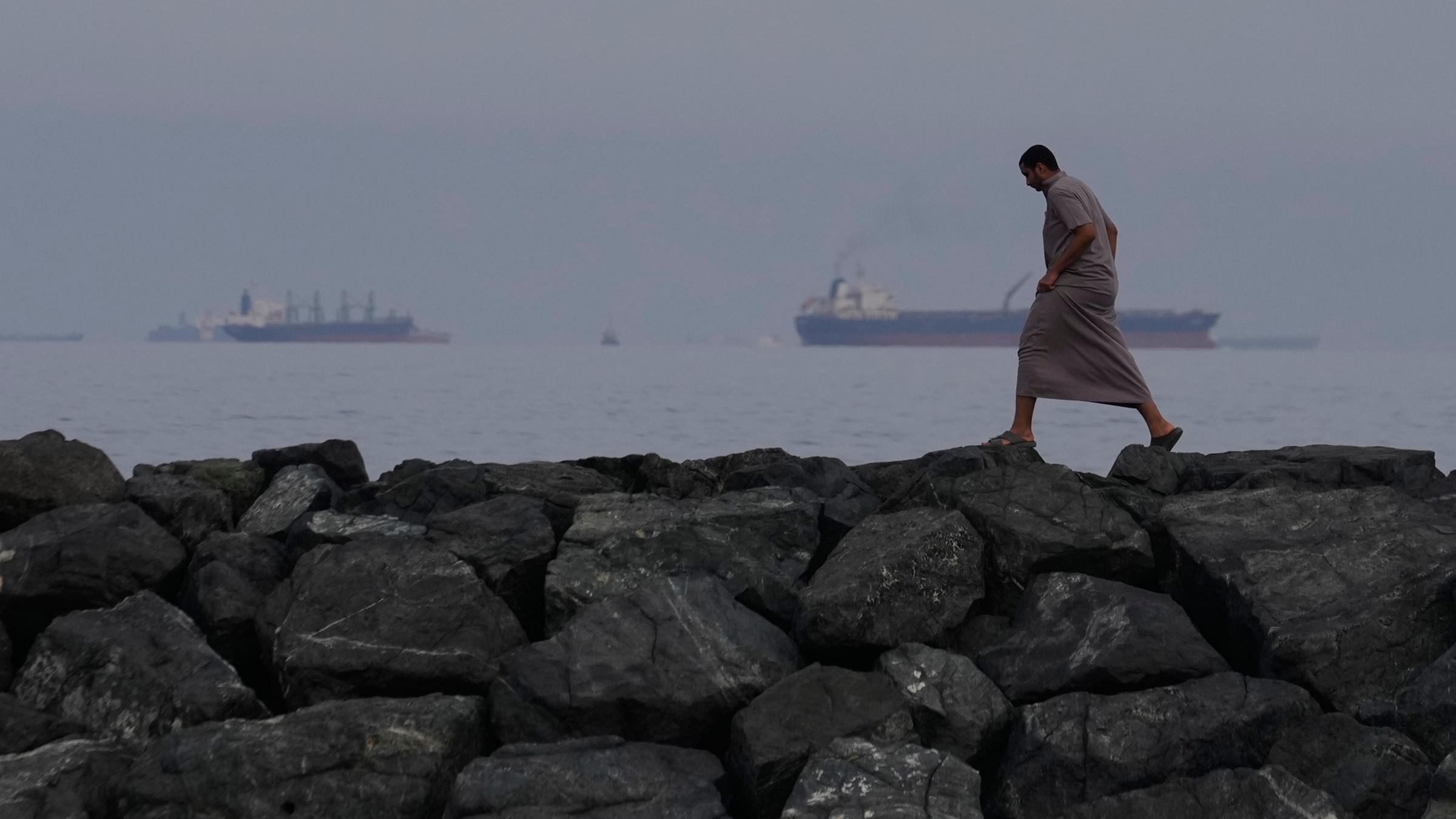 Ein Mann geht auf den Felsen am Ufer spazieren, während Öltanker und Frachtschiffe in der Straße von Hormus auf durchfahrt warten.